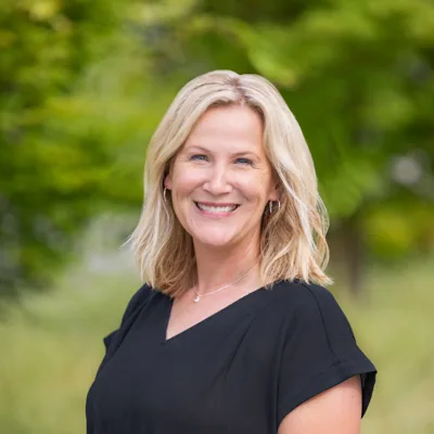 Headshot of Erin Toman smiling outdoors, with shoulder-length blonde hair, wearing a black top against a soft green, tree-filled background.