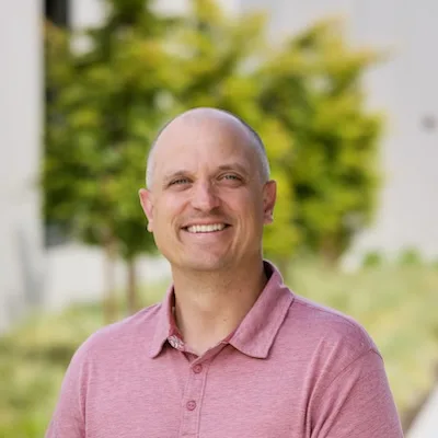 Headshot of Jason Irving, a smiling person with a shaved head wearing a pink polo shirt, outdoors with blurred greenery in the background.