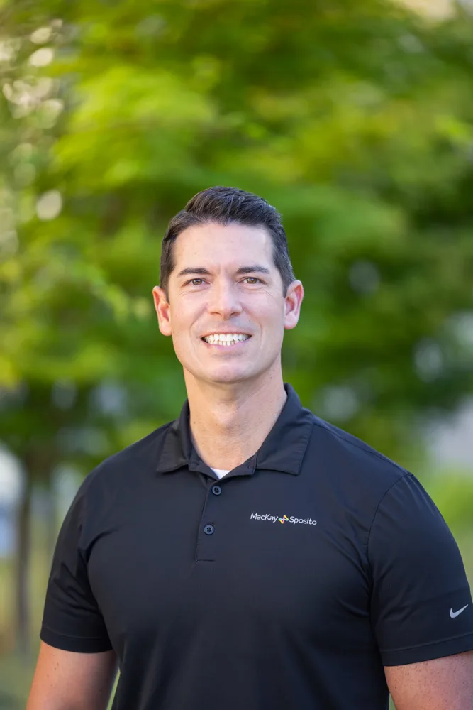 Headshot of Jason Joyce smiling in a black polo with the MacKay Sposito logo, outdoors against a blurred green background.