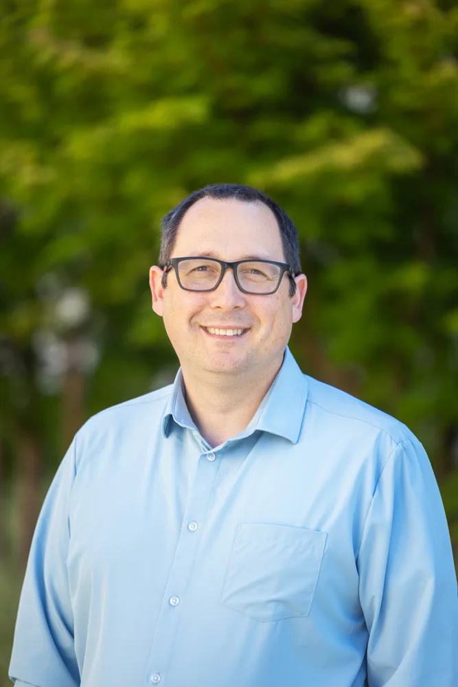 Headshot of Jerry Balch, a smiling person with short dark hair and glasses wearing a light blue button-down shirt against a blurred green foliage background.