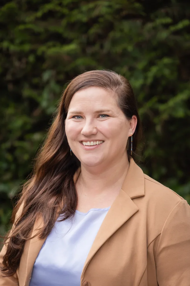 Headshot of Lisa Moore smiling outdoors, with long brown hair, wearing a tan blazer over a light blue top against a leafy green background.