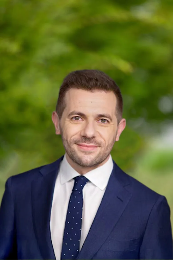 Headshot of Petru Santu Acquaviva, smiling in a navy suit, white shirt, and navy polka-dot tie against a blurred green outdoor background.