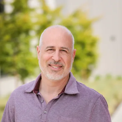 Headshot of Rob Palena smiling, with a shaved head and short gray beard, wearing a light purple polo against a softly blurred outdoor background with greenery.