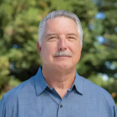 Older man with short gray hair and a mustache wearing a light blue collared shirt, standing outdoors with a blurred background of green trees.