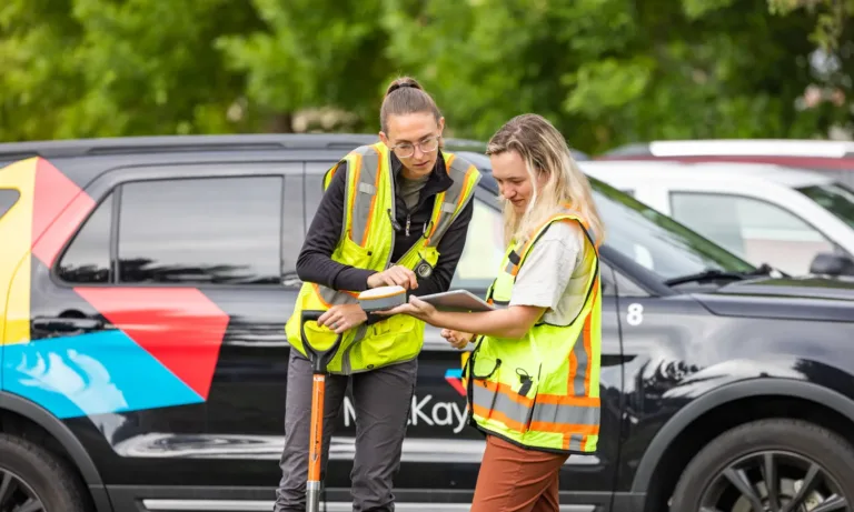 Two MacKay Sposito team members in high-visibility vests review plans on a tablet beside a branded SUV at a field site.