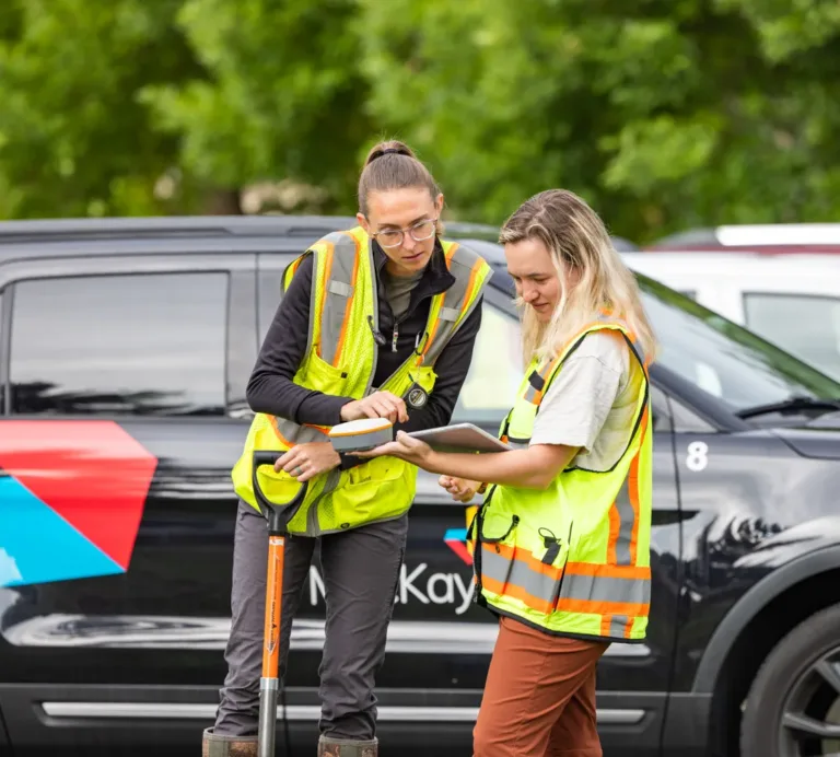 Two MacKay Sposito team members in high-visibility vests review plans on a tablet beside a branded SUV at a field site.