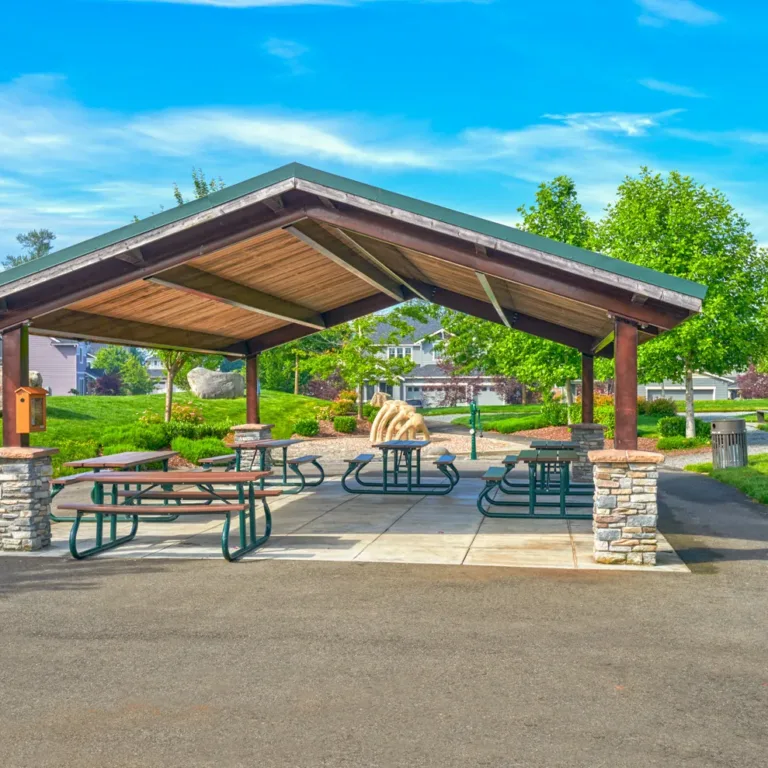 Open-air picnic pavilion with a wooden roof and stone columns, surrounded by green lawns and trees, with picnic tables and barbecue grills in a suburban park under a bright blue sky.