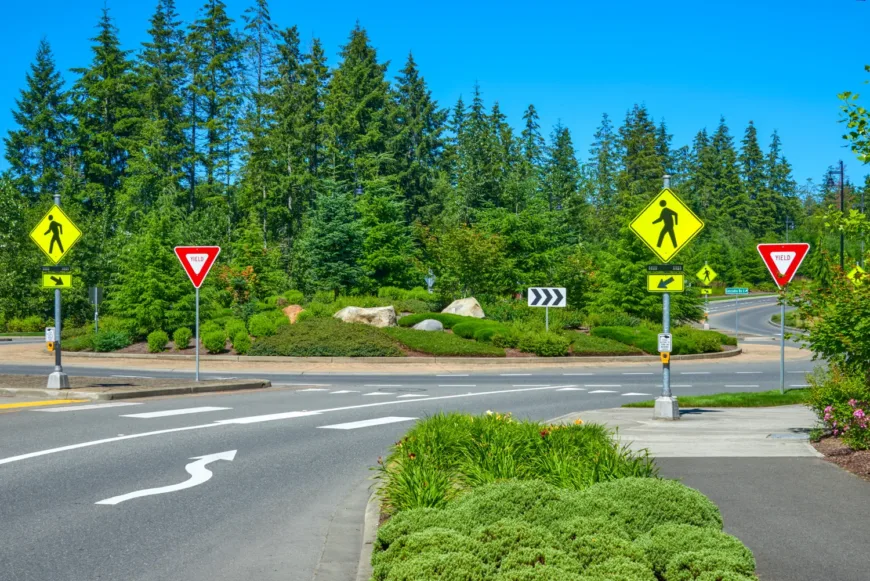 Landscaped roundabout with yield signs, pedestrian crossing signs and button, directional arrows on the roadway, and evergreen trees in the background under a clear blue sky.