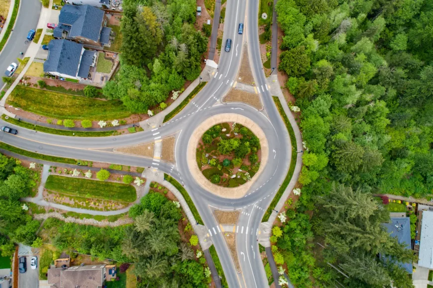 Aerial view of a single-lane three-way roundabout with a landscaped central island, marked crosswalks at each approach, and surrounding suburban homes and dense greenery.