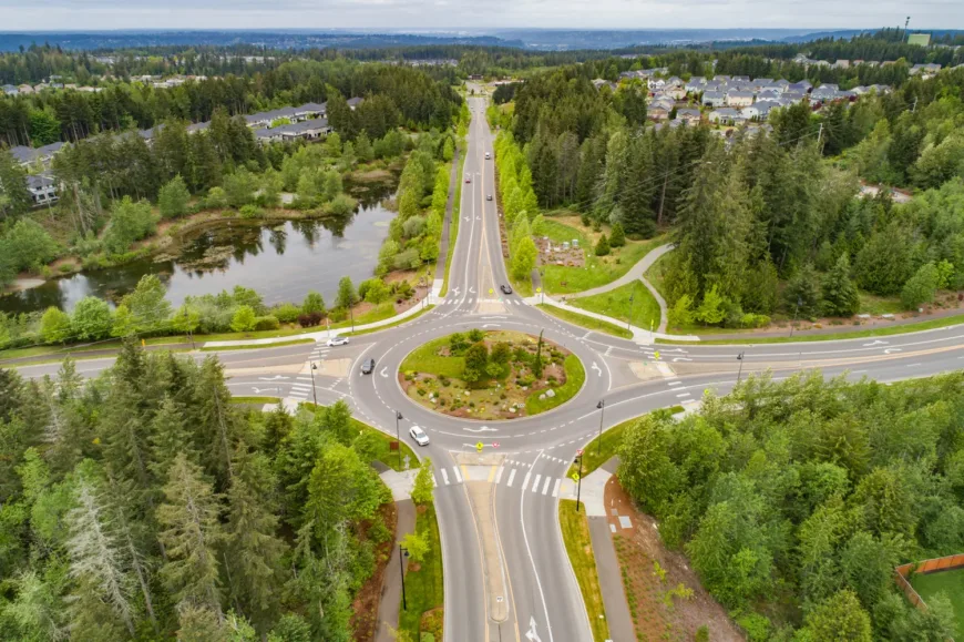 Aerial view of a landscaped multi-lane roundabout with crosswalks and a few cars, surrounded by evergreen trees, a small pond, and nearby suburban homes.