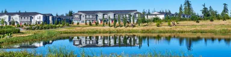Row of modern townhouses reflected in a calm pond, bordered by reeds and grass under a clear blue sky.
