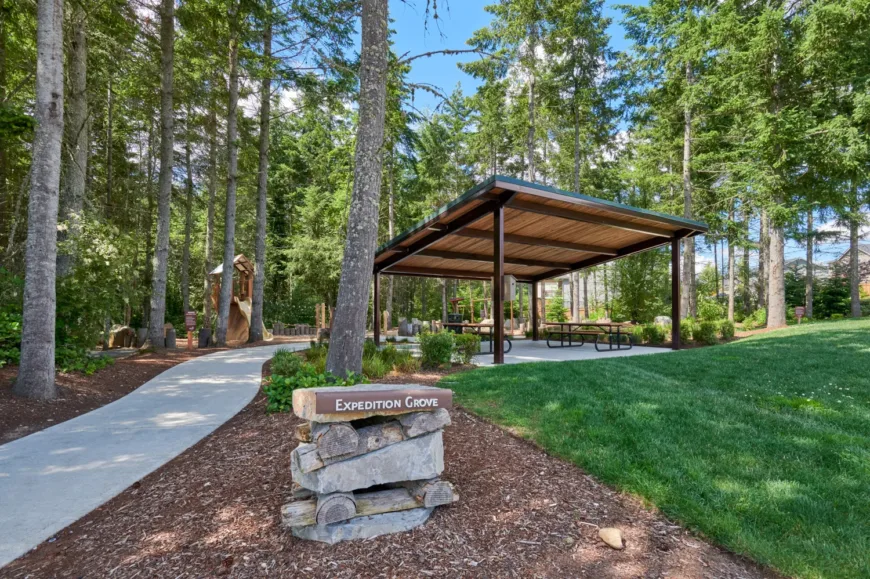 Shaded picnic pavilion with tables in a forested park labeled “Expedition Grove,” beside a curving path, a small play structure, and a grassy lawn under tall evergreens.