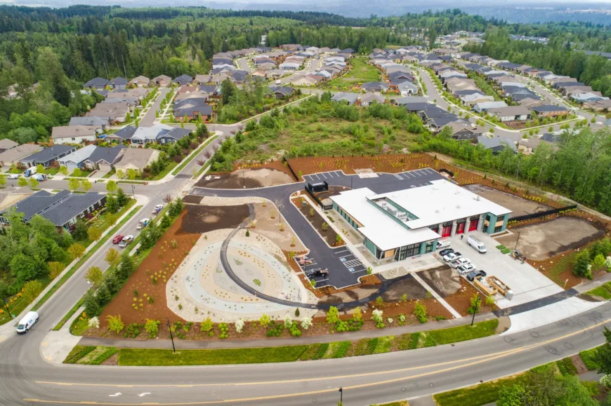 Aerial view of a suburban neighborhood bordered by forest, highlighting a modern single-story building with a white roof, red bay doors, parking lot, and landscaped grounds along curving streets.