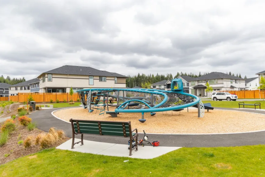 Modern blue climbing play structure with rope nets in a suburban park, with benches and maintenance tools in the foreground and houses under a cloudy sky in the background.