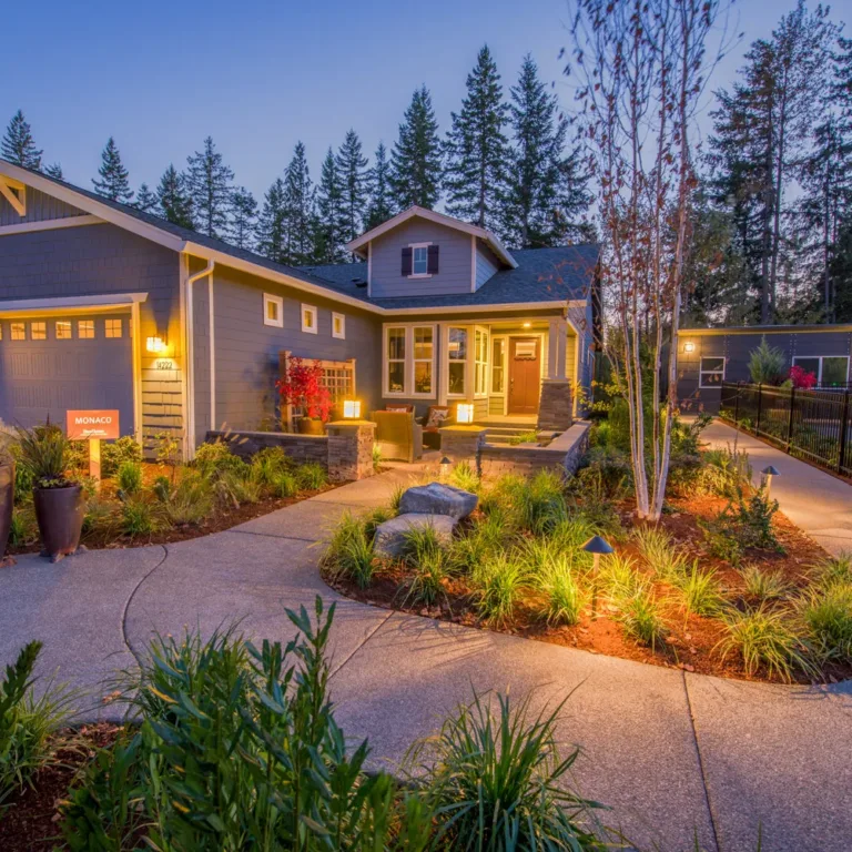 Craftsman-style gray single-story home at dusk with warm exterior lights, a curved concrete walkway through lush landscaped garden beds and potted plants, and tall evergreens in the background.