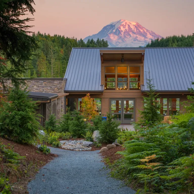 Modern wood-and-stone house with a steep metal roof and large windows, surrounded by lush evergreens along a gravel path, with a snow-capped mountain glowing at sunset in the background.