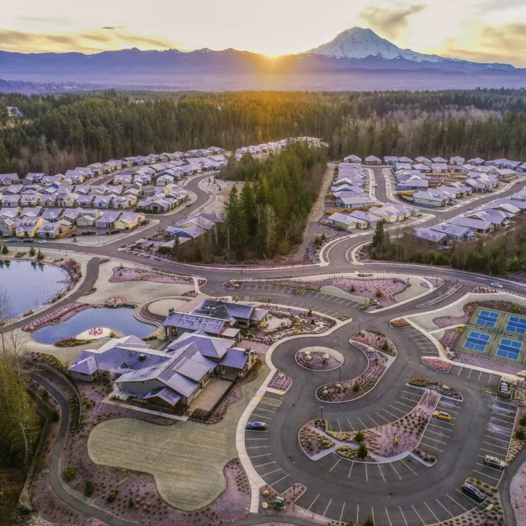 Aerial view of a large suburban community with a central clubhouse, ponds, parking lots, and blue pickleball/tennis courts, surrounded by winding roads and forest as the sun sets behind a distant snow‑capped mountain range.