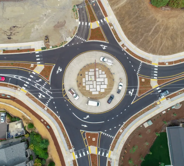 Aerial view of a newly paved roundabout with a geometric-tile central island, clear lane arrows and crosswalks, and several cars circulating through the intersection.