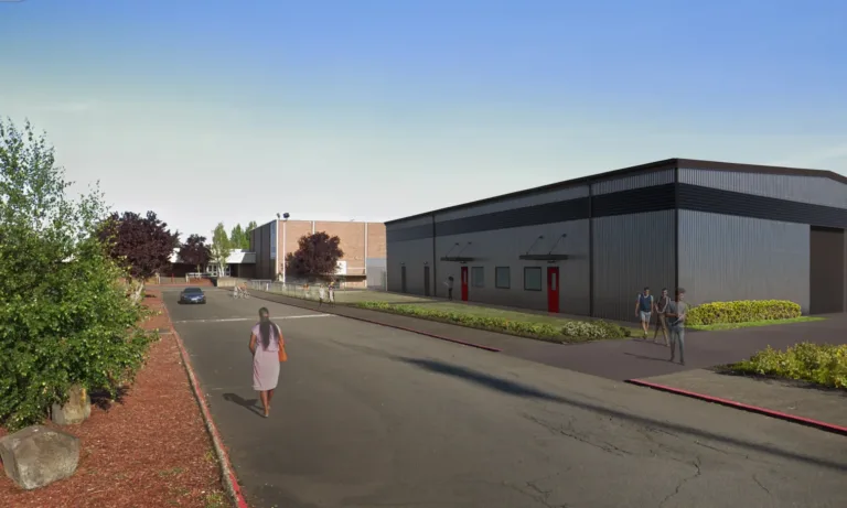 Corrugated metal building with black trim and red doors lines a campus road, with pedestrians, a car, and landscaped beds under a clear blue sky.