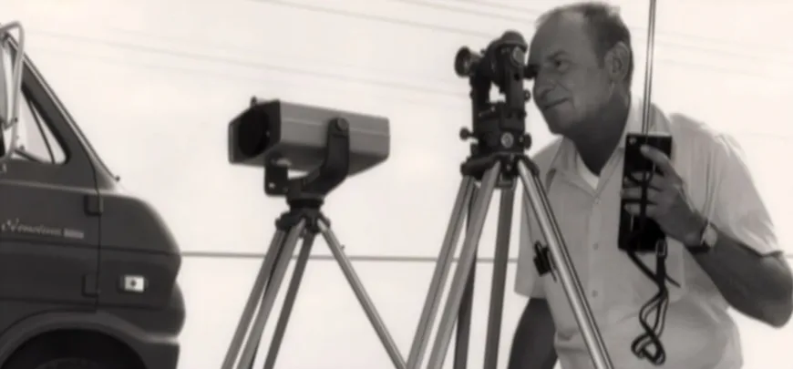 Historical MacKay Sposito photo of a surveyor operating tripod-mounted surveying instruments beside a van, in black and white.