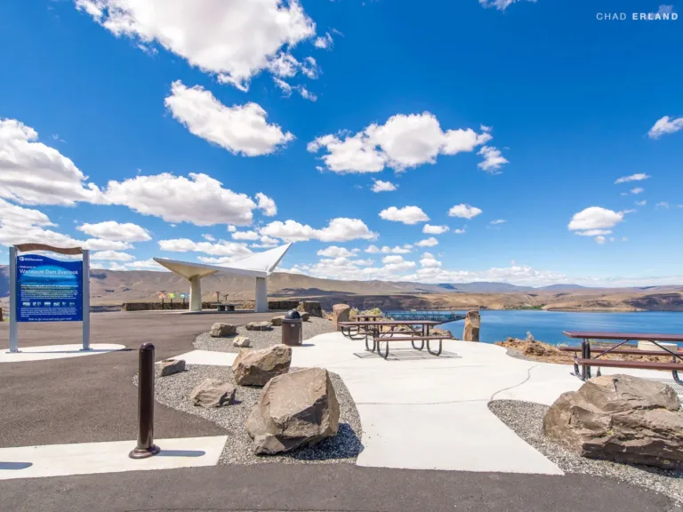 Wanapum Dam Overlook featuring picnic tables, boulders, and a modern white canopy, with sweeping views of a blue reservoir and arid hills under a bright, cloud-dotted sky.