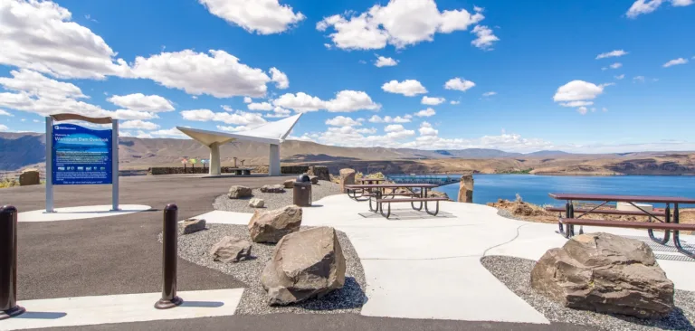 Wanapum Dam Overlook featuring picnic tables, boulders, and a modern white canopy, with sweeping views of a blue reservoir and arid hills under a bright, cloud-dotted sky.