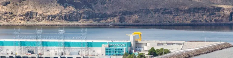 Wide view of Wanapum Dam spanning the Columbia River, with a long earthfill embankment and access road in the foreground, arid basalt hills behind, and a partly cloudy blue sky overhead.