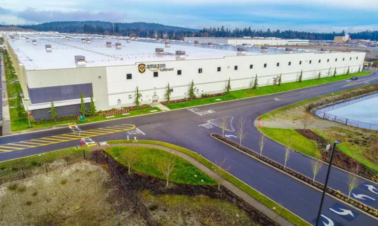 Aerial view of a large Amazon fulfillment center with a white flat‑roofed warehouse, landscaped driveways, and a drainage pond under an overcast sky.