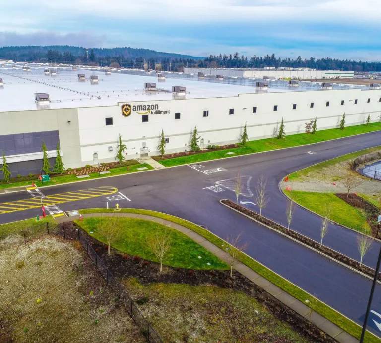 Aerial view of a large Amazon fulfillment center with a white flat‑roofed warehouse, landscaped driveways, and a drainage pond under an overcast sky.
