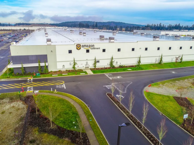 Aerial view of a vast Amazon Fulfillment warehouse with a white facade and flat roof, bordered by parking lots, landscaped greenery, and a curved access road.