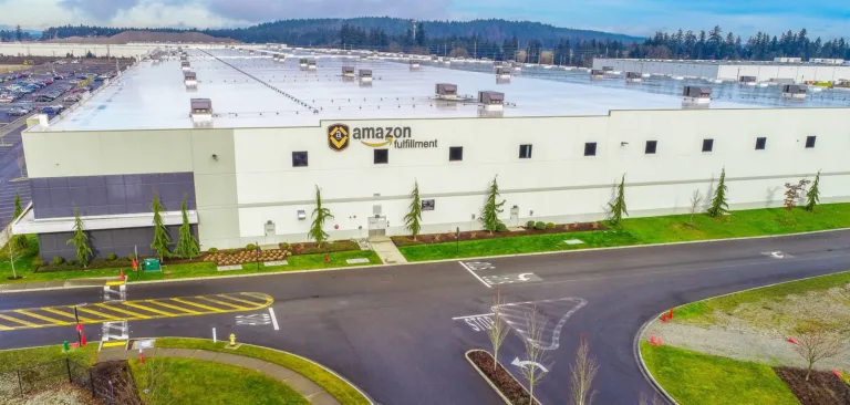 Aerial view of a vast Amazon Fulfillment warehouse with a white facade and flat roof, bordered by parking lots, landscaped greenery, and a curved access road.