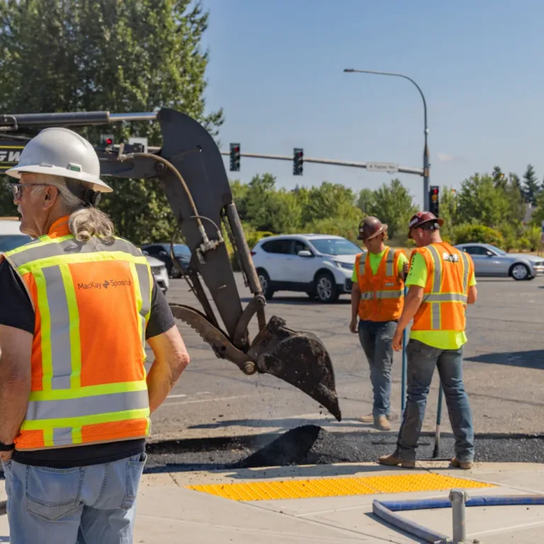 MacKay Sposito field crew in high-visibility vests oversee curb-ramp roadwork at a busy intersection with an excavator.