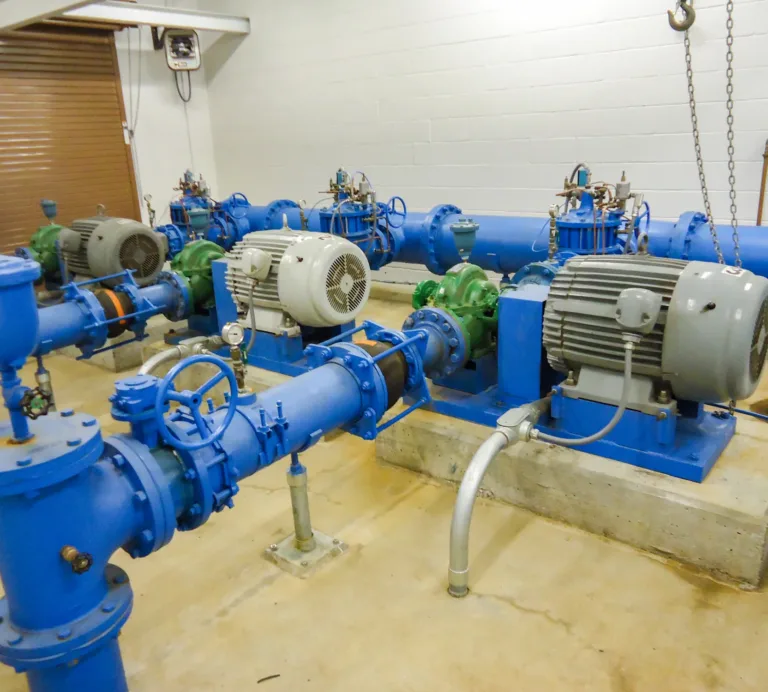 Industrial pump room with multiple blue pipes, valves, and gauges connected to large electric motors driving centrifugal pumps mounted on concrete pads.