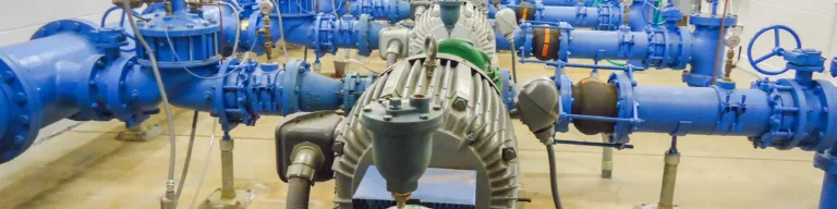 Industrial pump room with large blue pipelines, gauges and valves, a green centrifugal pump in the foreground, and a technician by electrical control panels in the background.