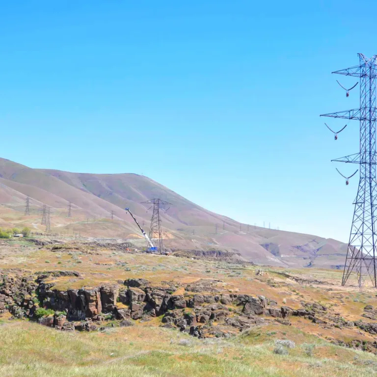 High-voltage transmission tower stands in a dry, rocky plain with rolling hills under a clear blue sky, with additional pylons and a crane assembling another tower in the distance.