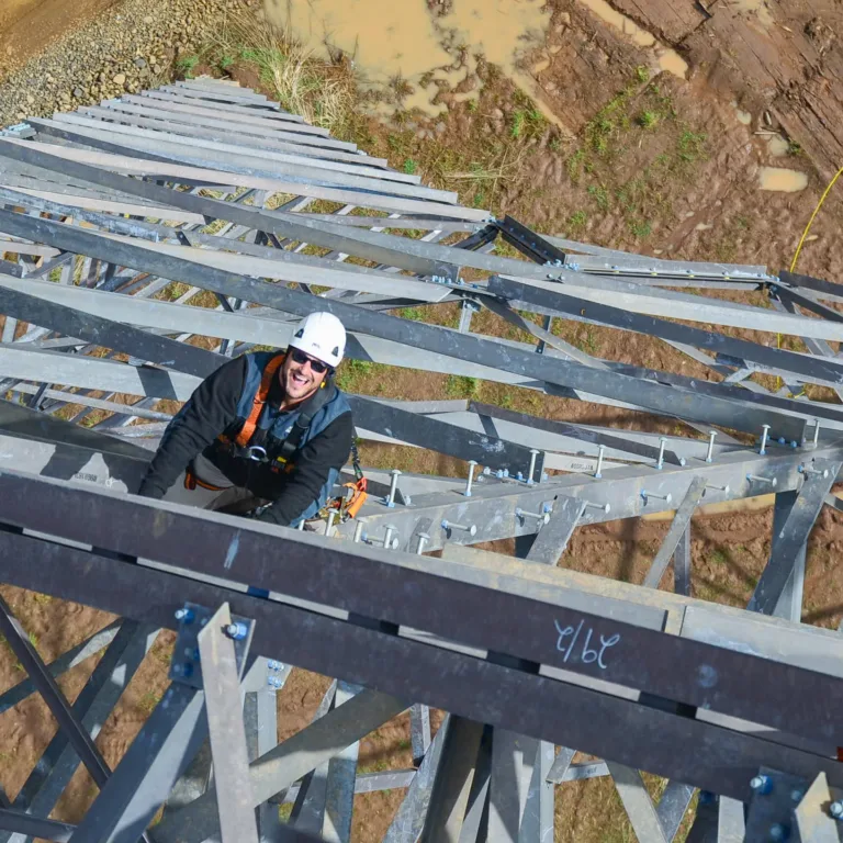 Worker in a white hard hat and safety harness climbs a steel lattice tower, seen from above with muddy ground below and beams and bolts forming a geometric pattern.
