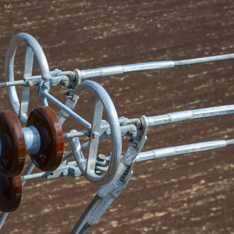Close-up of high-voltage transmission hardware showing brown ceramic disc insulators and an aluminum spacer joining three bundled steel conductors against a blurred earth-toned background.