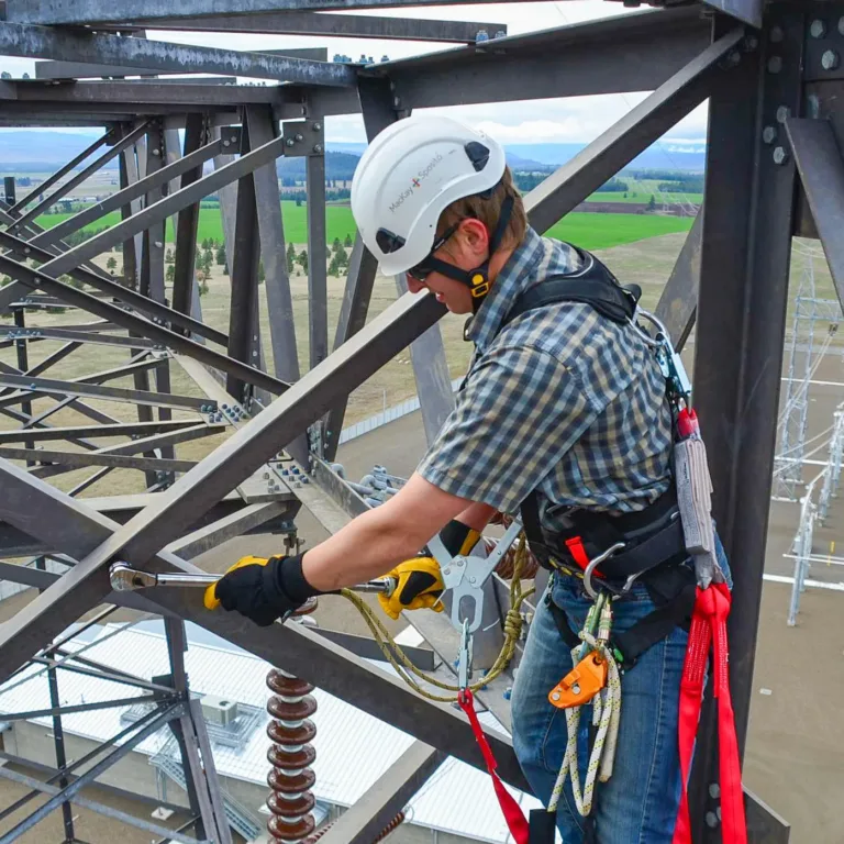 Utility worker in a helmet and safety harness tightens bolts with a ratchet on a high steel transmission tower, secured by lanyards above an electrical substation and surrounding fields.