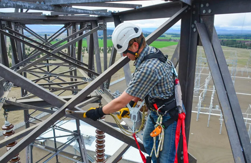 Utility worker in a helmet and safety harness tightens bolts with a ratchet on a high steel transmission tower, secured by lanyards above an electrical substation and surrounding fields.