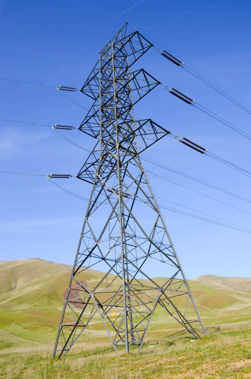 A tall steel high‑voltage transmission tower with power lines stands over rolling grassy hills under a clear blue sky.