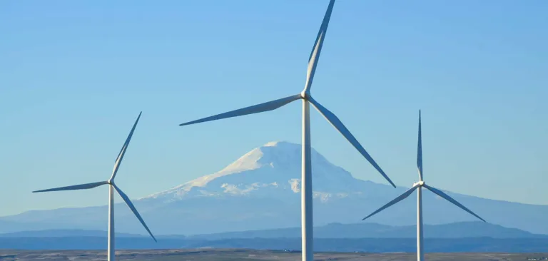 Three white wind turbines on rolling plains with a snow-capped mountain in the background beneath a clear blue sky.
