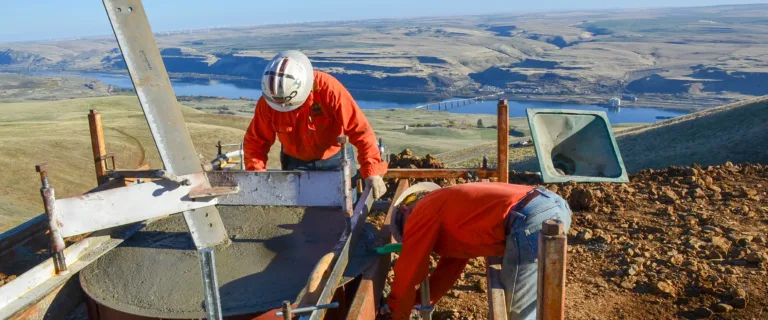 Two construction workers in orange safety gear and hard hats pour and level concrete in a cylindrical foundation form on a hillside, with scaffolding around them and a river valley and bridge in the background.