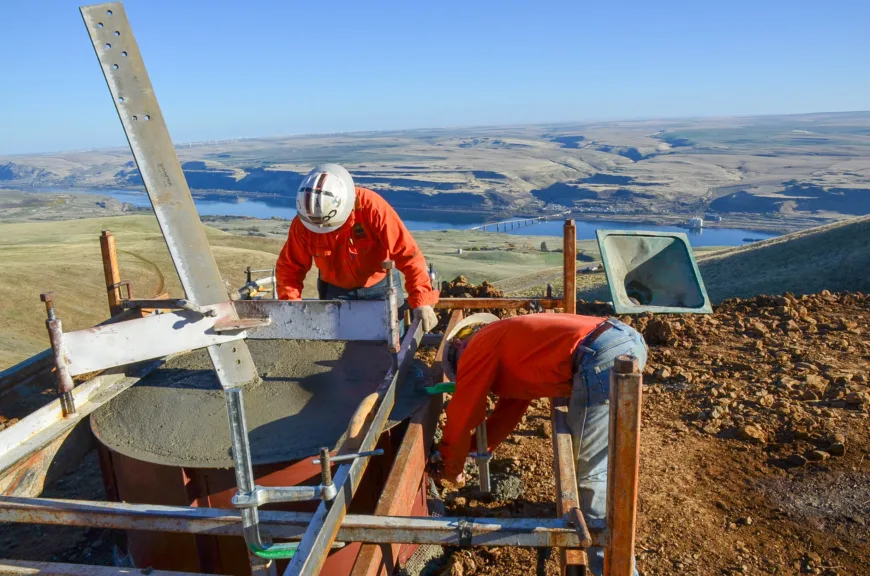 Two construction workers in orange safety gear and hard hats pour and level concrete in a cylindrical foundation form on a hillside, with scaffolding around them and a river valley and bridge in the background.