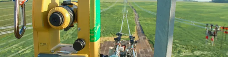 Yellow total station mounted on a transmission tower, overlooking high‑voltage power lines and glass insulators stretching across green fields.