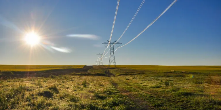 High-voltage transmission towers and lines span a grassy rural plain beneath a bright, low sun and a clear blue sky.