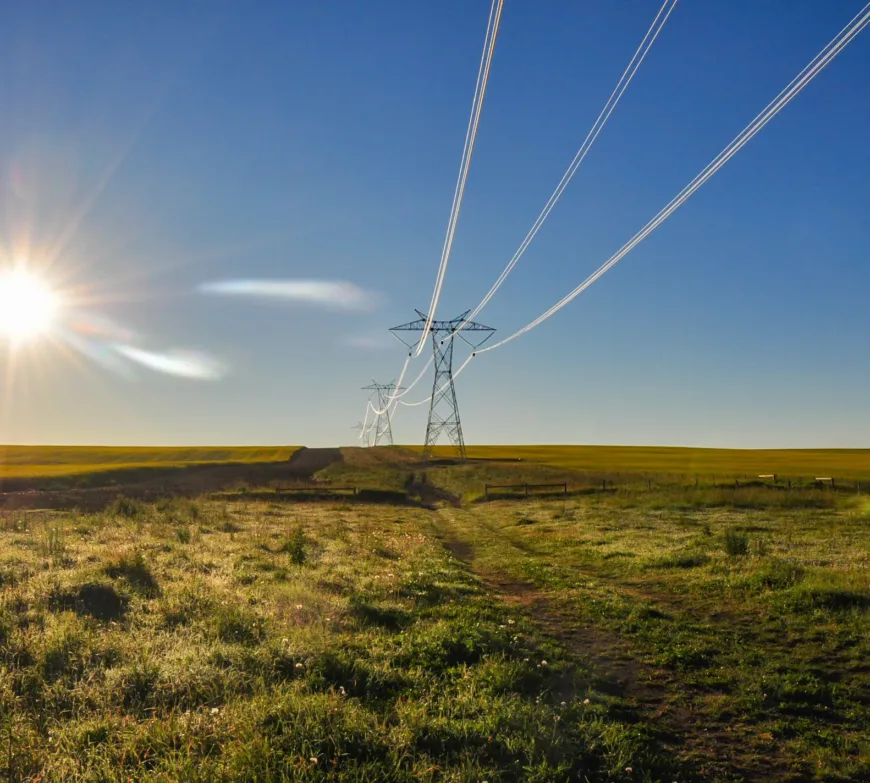 High-voltage transmission towers and lines span a grassy rural plain beneath a bright, low sun and a clear blue sky.