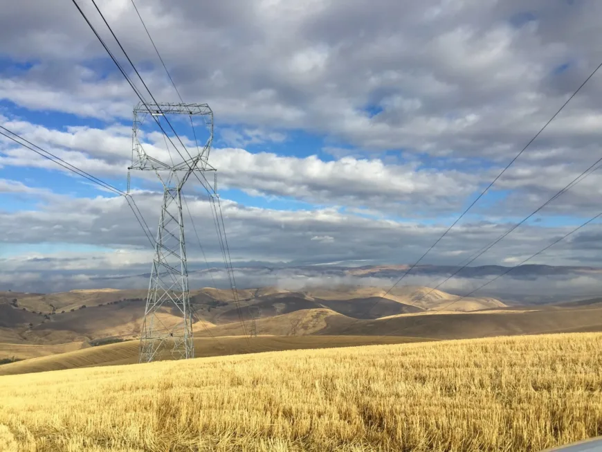 A steel electricity transmission tower with power lines crossing a golden harvested field, with rolling hills and a partly cloudy blue sky in the background.