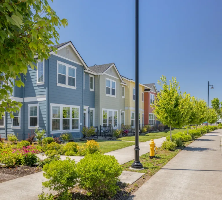 Row of colorful modern townhouses with landscaped gardens along a tree-lined sidewalk and parked cars on a sunny suburban street.