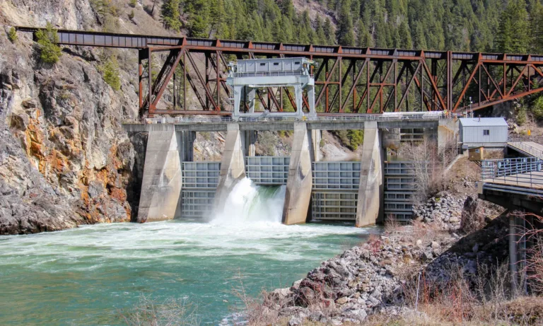 Concrete dam with a spillway releasing water beneath a rusted steel truss railway bridge in a forested mountain gorge.