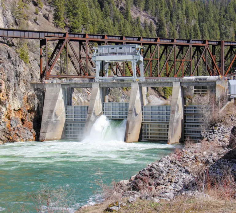 Concrete dam with a spillway releasing water beneath a rusted steel truss railway bridge in a forested mountain gorge.